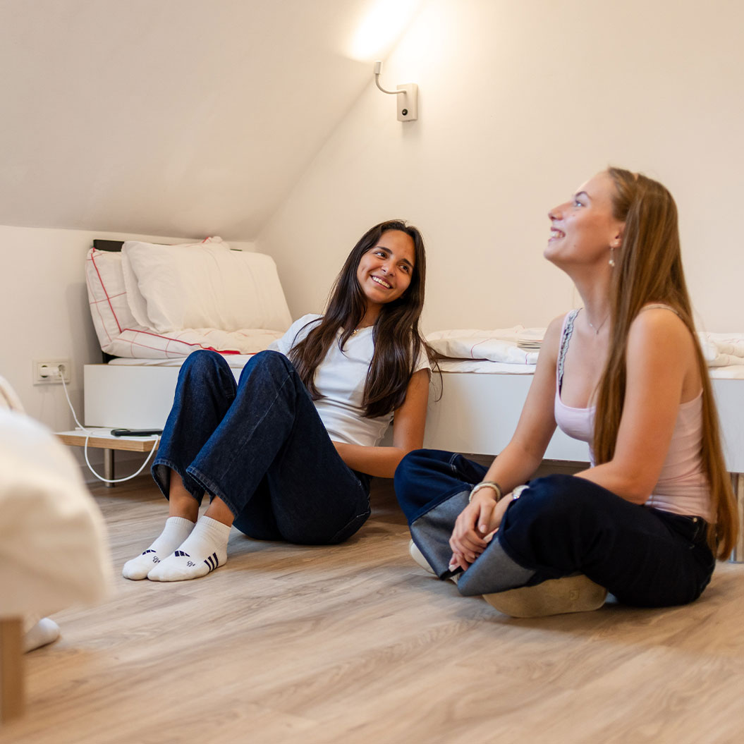 Zwei lächelnde Frauen in Weiß und Rosa sitzen auf Holzboden im Zimmer