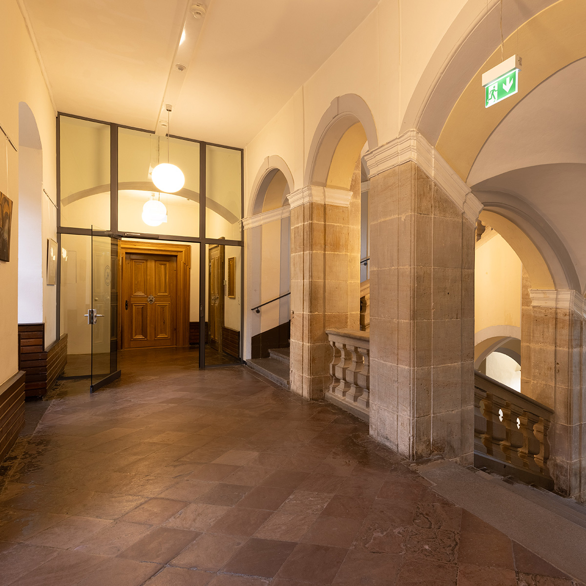 Leerer Flur mit Steinboegen und Holztaer, warmes Licht, Notausgangschild