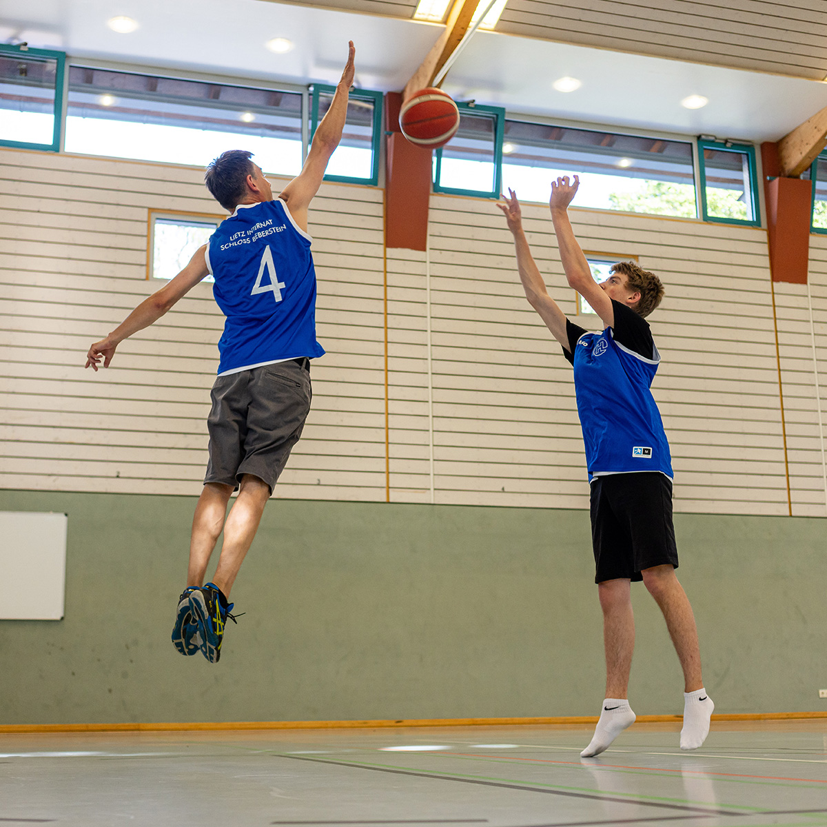 Zwei Spieler in blauen Trikots, konzentriert beim Basketballwurf in heller Halle