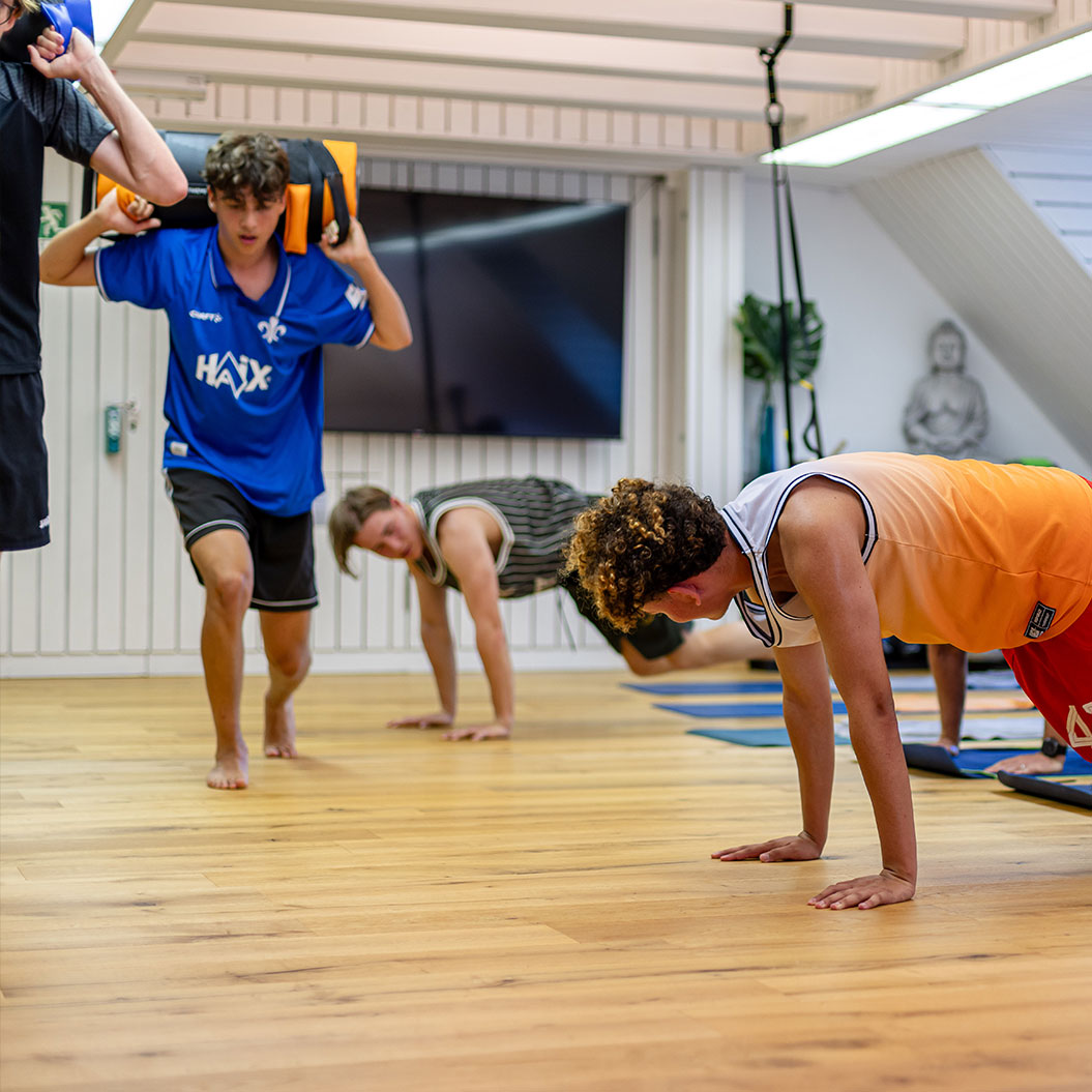 Junge in blauem Trikot schwitzt konzentriert, andere in Plank, bunte Matten