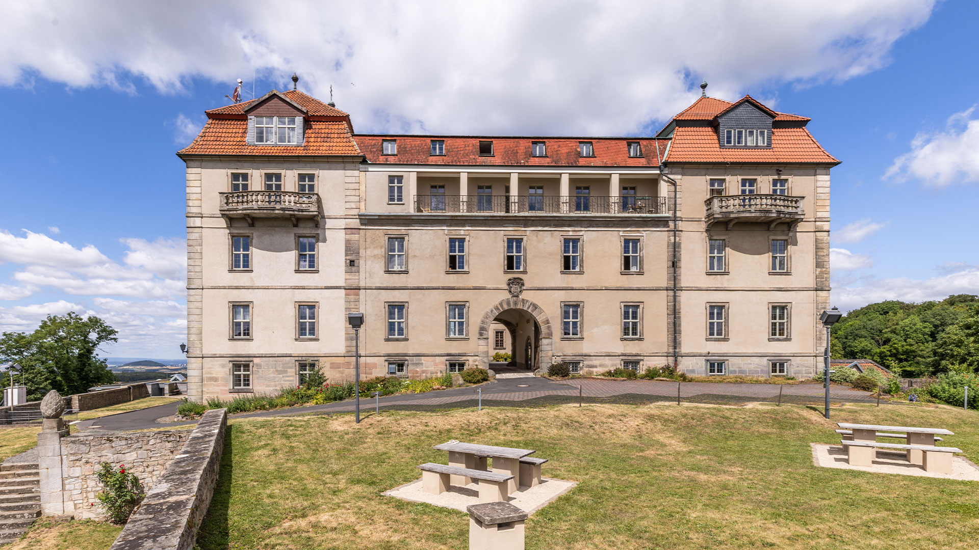 Beiges Schloss mit rotem Dach und Torbogen vor gruenem Hof und Himmel
